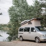 A silver Volkswagen campervan with pop-top roof parked beside a lake, string lights hanging from the trees