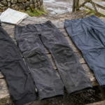 Three pairs of walking trousers laid out with hiking boots and a map on a trail bench
