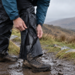 Hiker pulling on black waterproof overtrousers over muddy walking boots in wet UK conditions